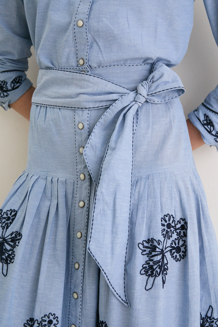 The Chambray Embroidered Highland Skirt features a classic, effortless design with floral embroidery, a self-tie sash, pleats, and fun snap buttons for a high-end look.