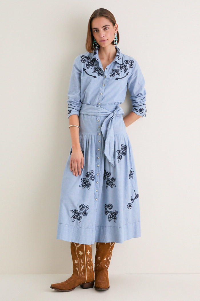 Young woman in a powder-blue shirtdress with black embroidery, showcasing The Chambray Embroidered Highland Skirt's classic, effortless style, paired with embroidered cowboy boots and turquoise earrings.