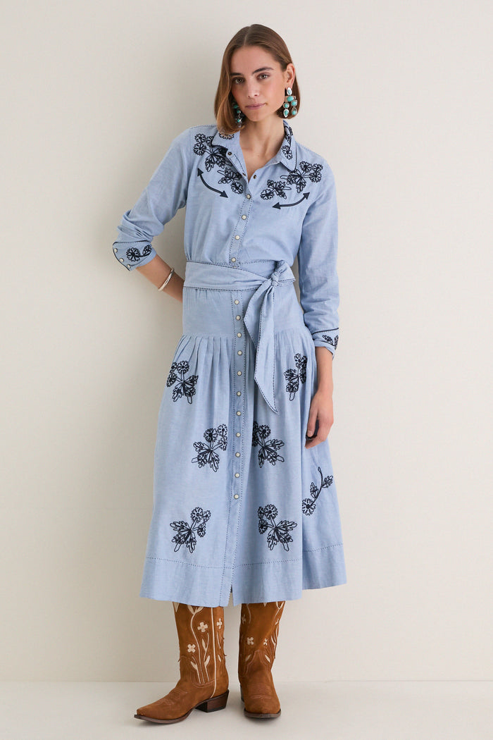 Woman models The Chambray Embroidered Western Shirt with floral embroidery, rolled sleeves, and a self-fabric belt, paired with turquoise earrings and cowboy boots, embodying classic, effortless style.