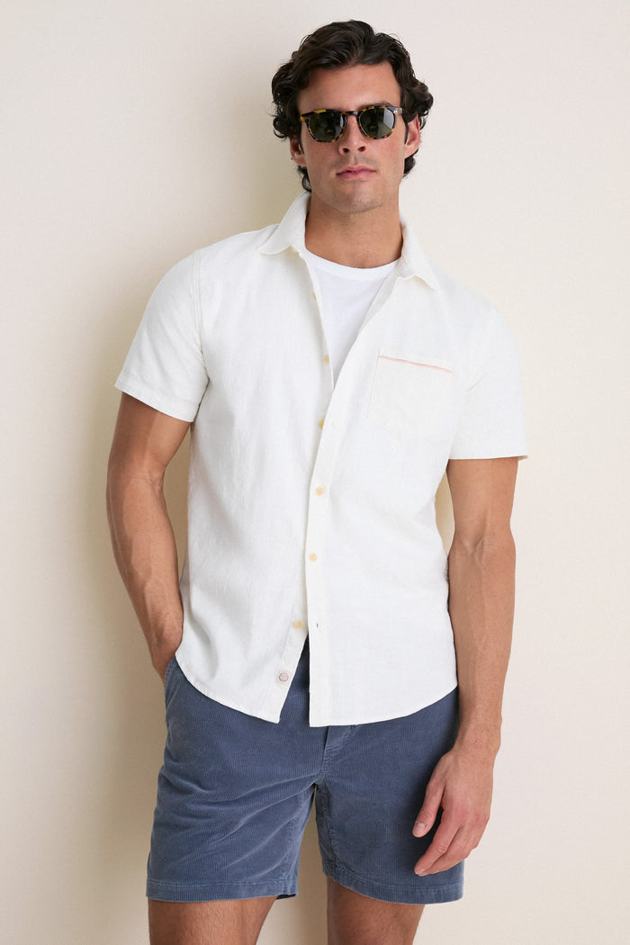 Man in an effortless, high-end summer look featuring a classic white stretch selvage shirt by Marine Layer, worn open over a T-shirt with blue shorts.