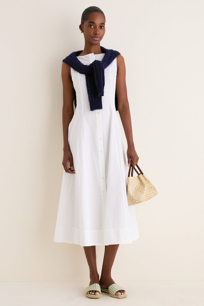 Young woman in a classic, sleeveless White Cotton Boat Neck Dress, draped with a navy sweater, carrying a straw tote, embodying an effortless, high-end resort style.