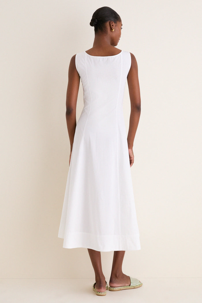 Back view of woman in white cotton boat neck dress, fitted bodice, A-line skirt. Hair in a bun, gold hoops, slip-on sandals. Classic, effortless style.
