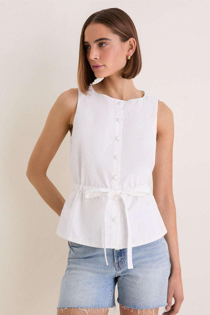 Young woman in a classic, sleeveless White Cotton Poplin Francoise Top with a high neckline and self-tie waist, paired with light-wash denim shorts, exuding effortless elegance.