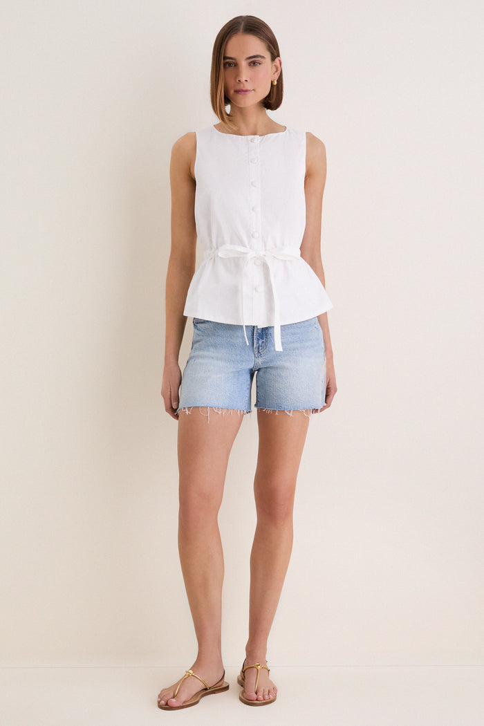 Young woman in classic, high-end White Cotton Poplin Francoise Top with chic high neckline and self-tie belt, paired with light denim shorts and gold-tone sandals. Effortless style.