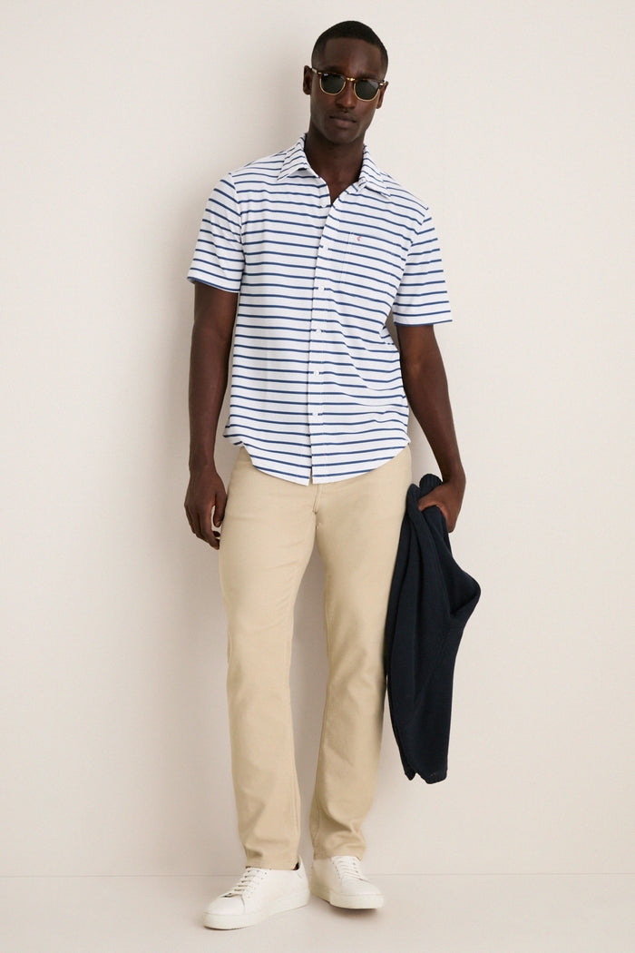 Man wearing the classic White Performance Pique Knit Button Down with navy stripes, khakis, and sneakers, holding a navy jacket. Effortlessly high-end and perfect for fun, warm days.