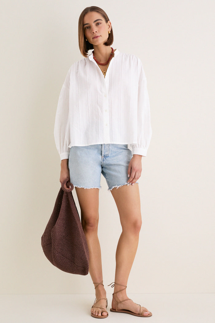 Woman models an effortless, high-end White Shadow Stripe Juliette Button Down with a classic flowy silhouette, paired with frayed denim shorts and strappy sandals.