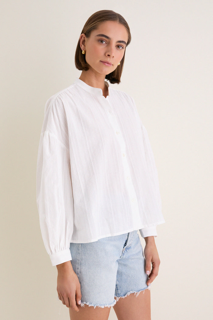 White Shadow Stripe Juliette Button Down embodies classic, effortless style with gathered sleeves and an asymmetrical hem, paired with light-wash frayed-hem denim shorts for a fun, high-end look.