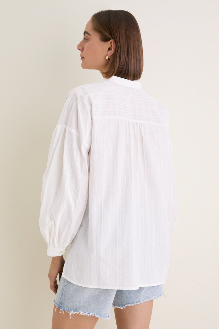White Shadow Stripe Juliette Button Down with flowy silhouette, gathered sleeves, and asymmetrical hem, worn with denim shorts. Effortless classic style for a fun, high-end look.