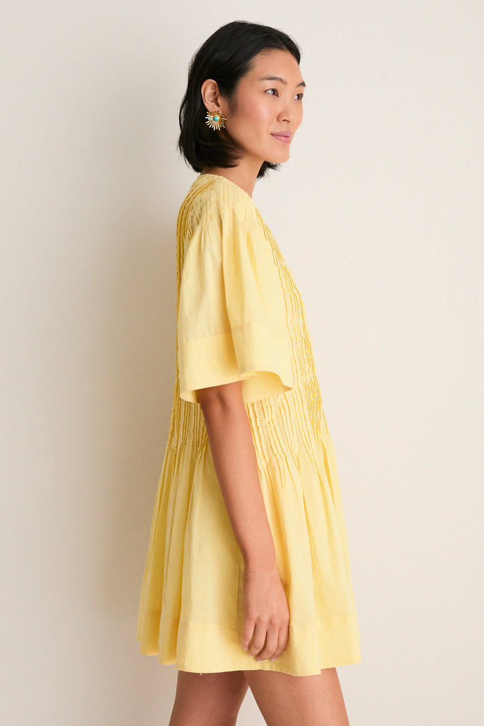Young woman in profile wearing the Yellow Alberte Solid Mini Dress, showcasing its classic, effortless style with pintuck pleating and sunburst earrings, embodying fun, high-end fashion.