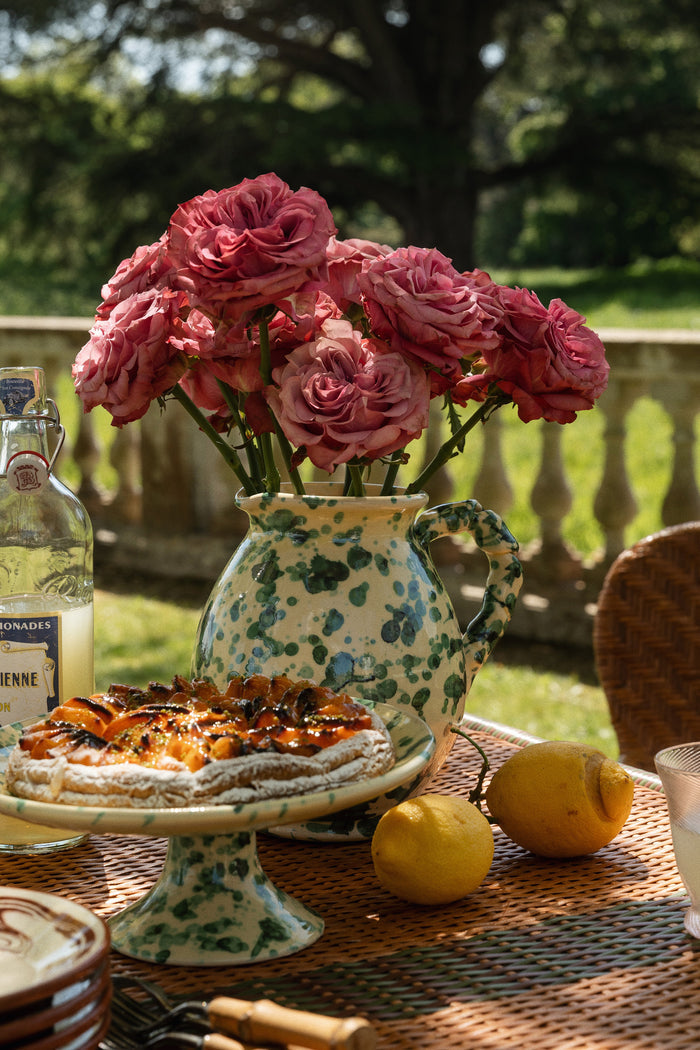 Yellow and Verde Splatter Cigno Jug, a classic ceramic pitcher, filled with pink roses, epitomizes effortless elegance on a high-end outdoor table setting.