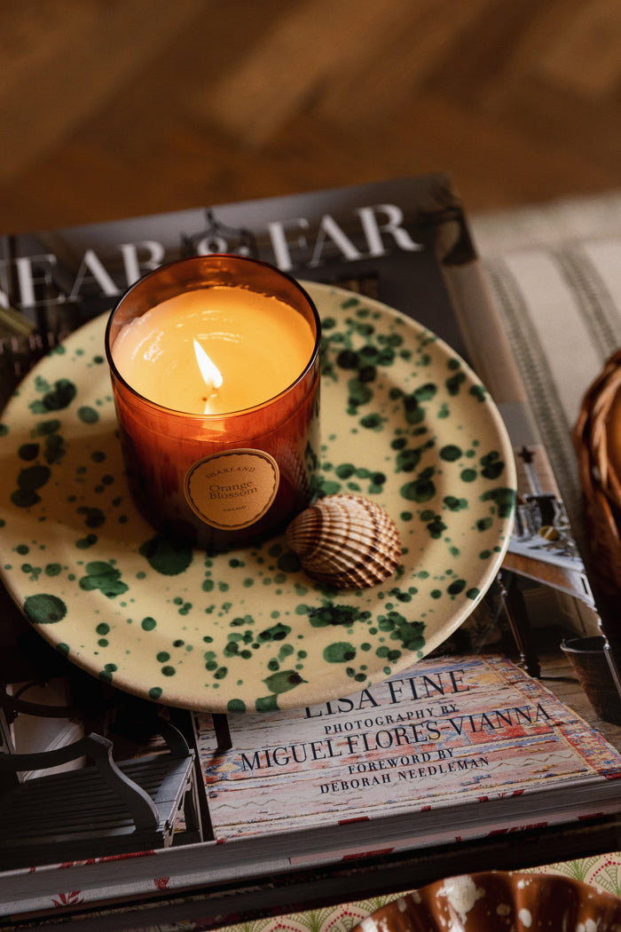 Amber candle on a ceramic Yellow and Verde Splatter Side Plate, paired with a seashell, atop Lisa Fine coffee-table book. Classic, high-end design for effortless, fun entertaining.
