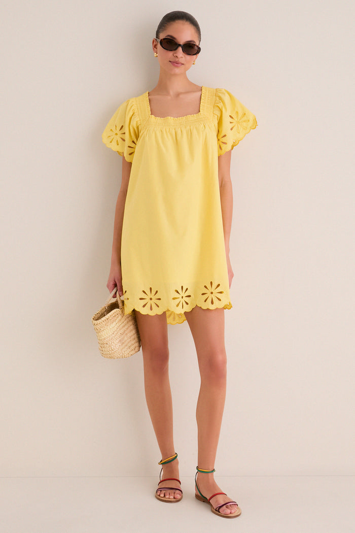 Woman in a classic, yellow Floral Eyelet Daisy Mini Dress with puffed sleeves, scalloped hem, paired with cat-eye sunglasses and a straw bag, exuding effortless, fun style.